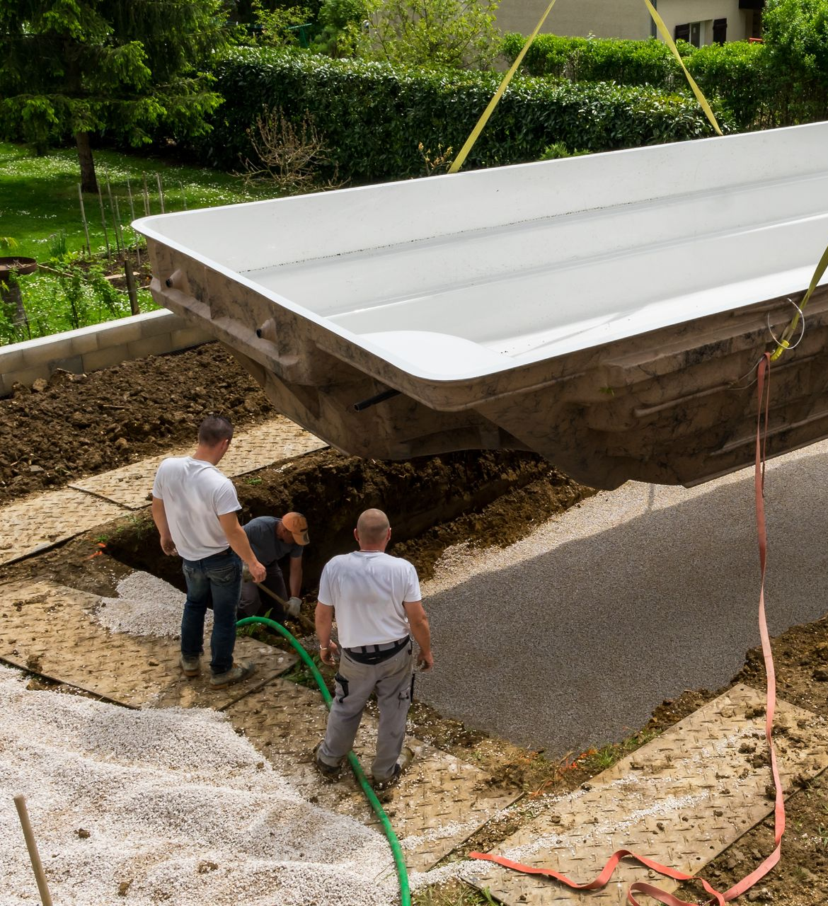 Trois hommes installent une piscine en fibre de verre blanche, la descendant dans une zone excavée à l'aide d'une grue.