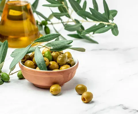Cuenco de aceitunas verdes, aceite de oliva y rama de olivo sobre mármol.