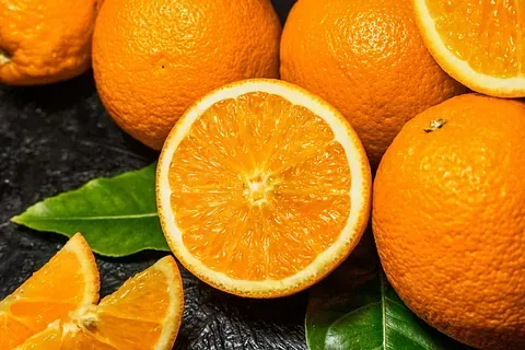 Primer plano de naranjas, incluyendo una cortada por la mitad, con hojas verdes. Fruto de color naranja brillante sobre una superficie oscura.