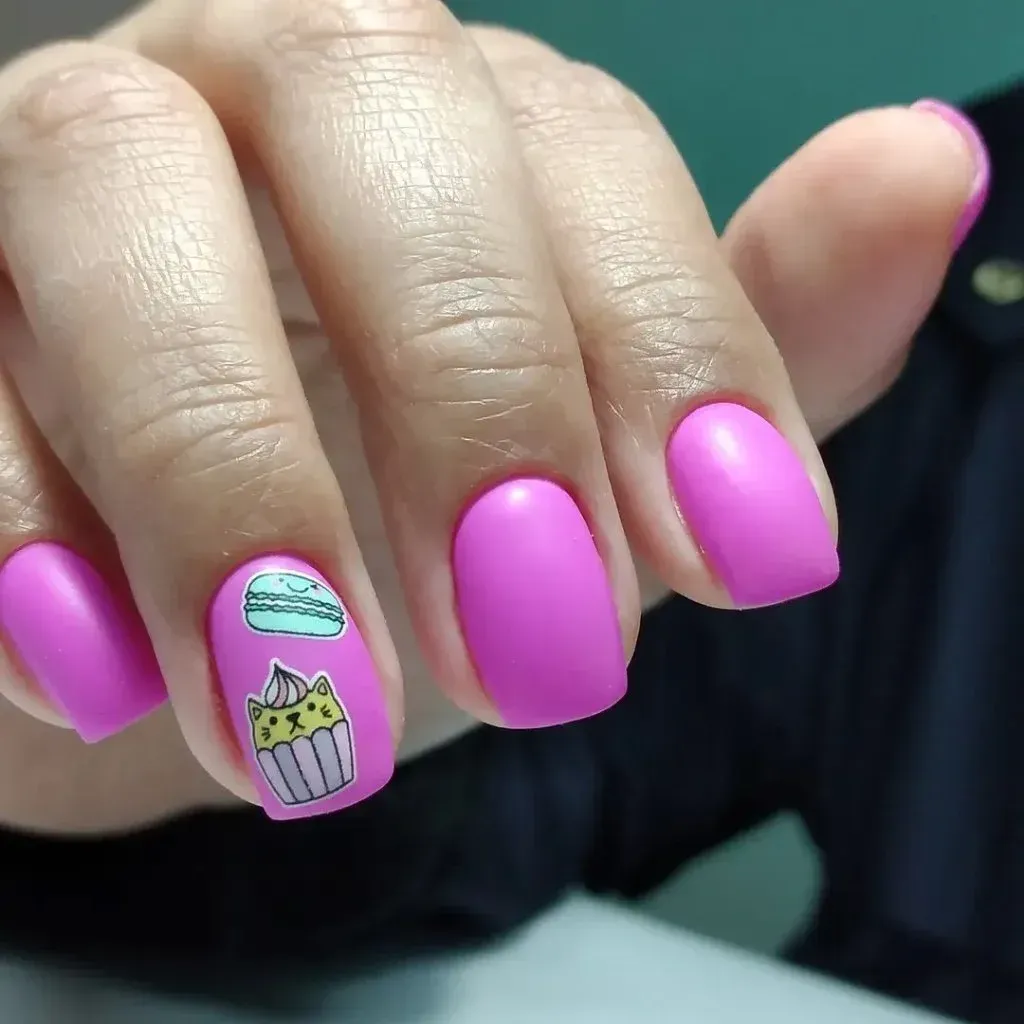 Mano con uñas de color rosa brillante y un diseño de arte de uñas de cupcake sobre un fondo verde.