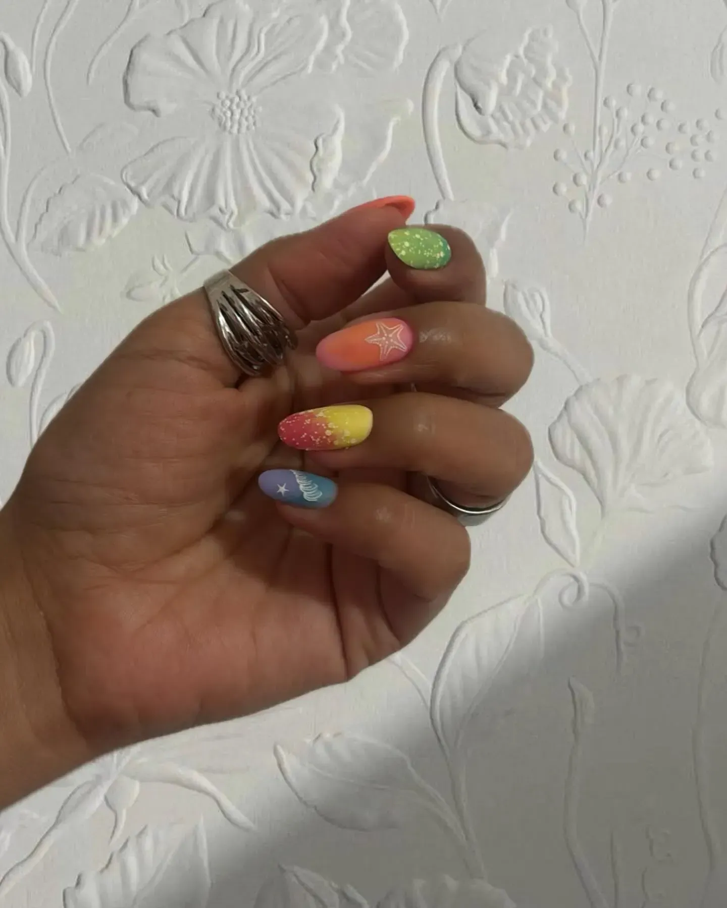 Mano con uñas de purpurina de colores pastel sobre una pared blanca texturizada.