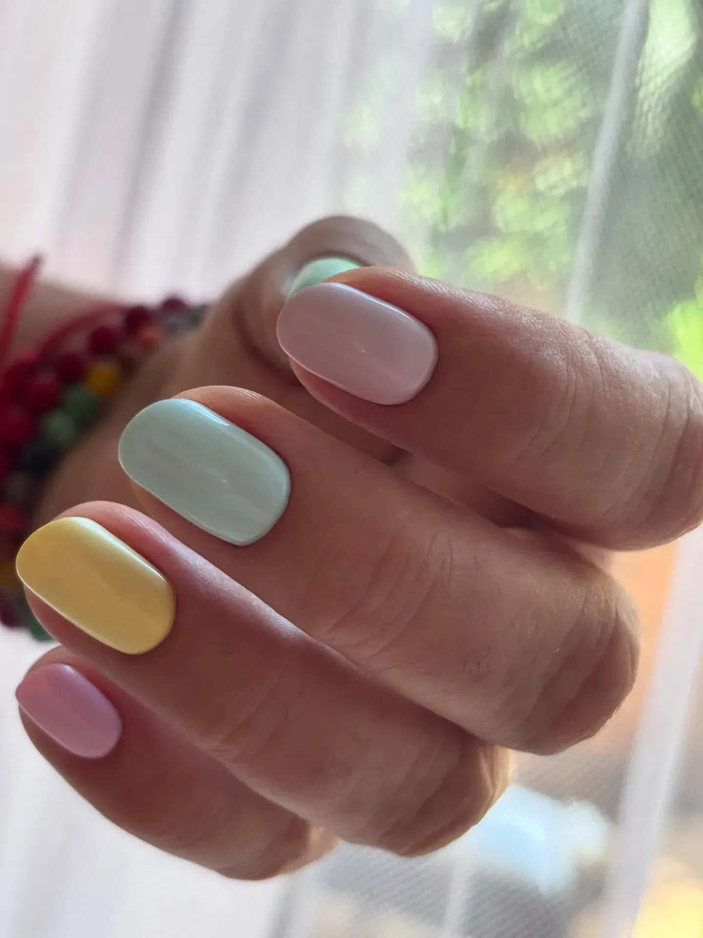 Mano con uñas manicuradas en tonos rosa pastel, menta, amarillo y rosa, sostenida cerca de una ventana suavemente borrosa.