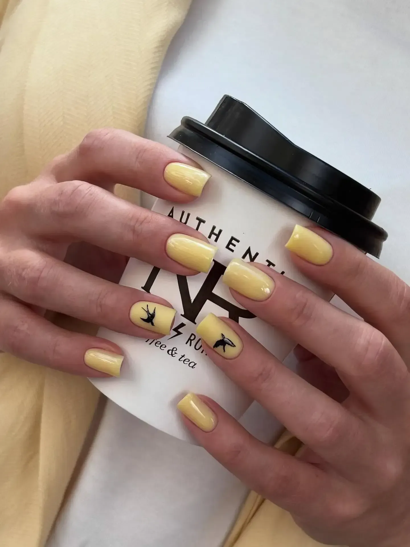 Manos con uñas amarillas sosteniendo una taza de café blanca con tapa y logotipo negros.