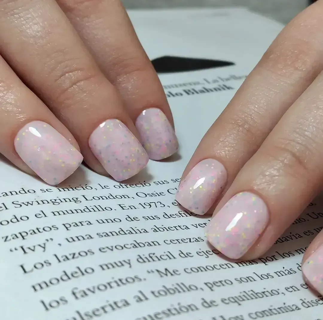 Manos con uñas de color rosa pálido con purpurina descansando sobre la página de un libro abierto.