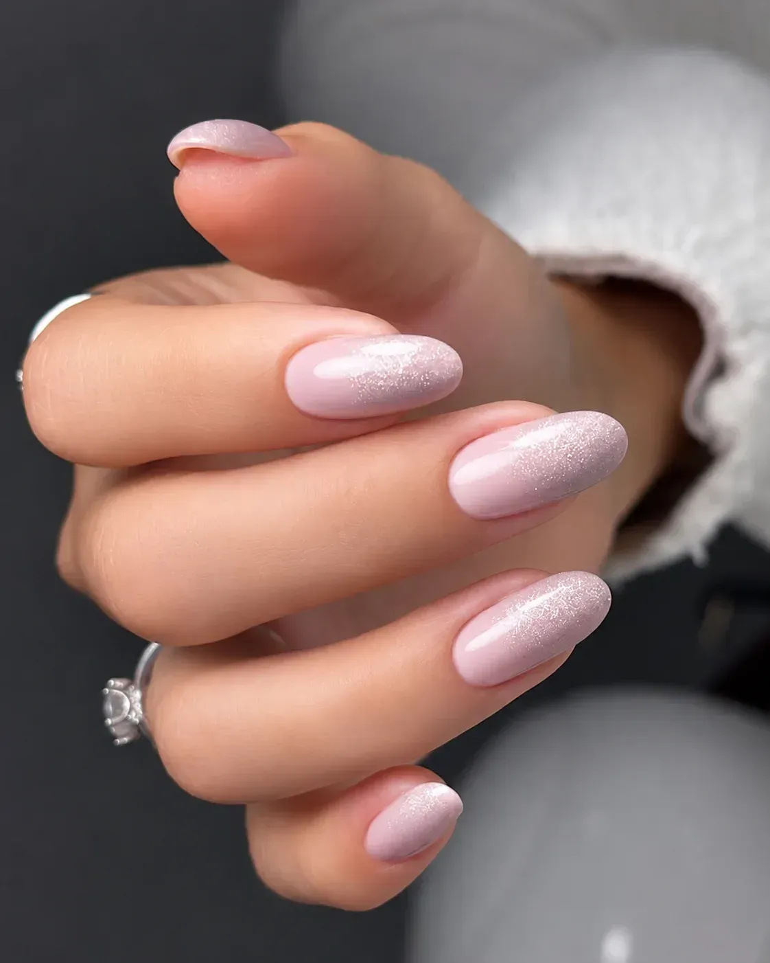 Mano con uñas largas de color rosa pálido con purpurina, mostrando un anillo sobre un fondo gris suave.