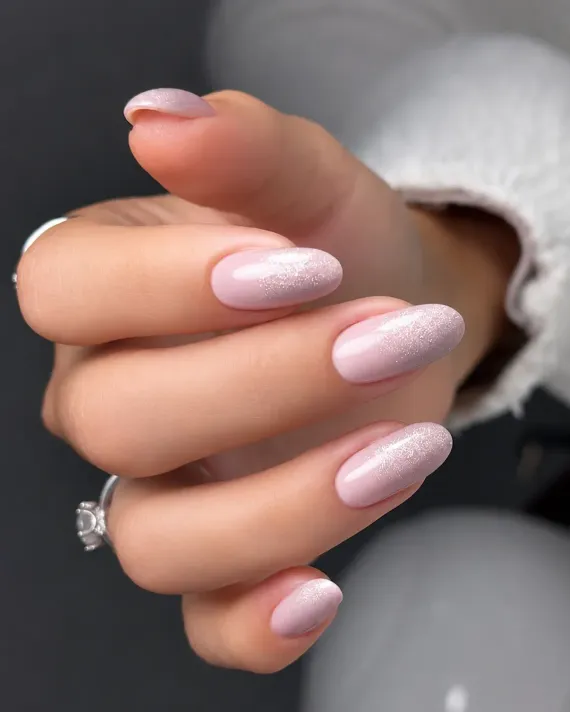Mano con uñas largas de color rosa pálido con purpurina, mostrando un anillo sobre un fondo gris suave.