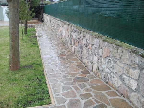 Sendero de piedra junto a un muro de piedra y una valla verde, con césped y árboles a lo largo del camino.