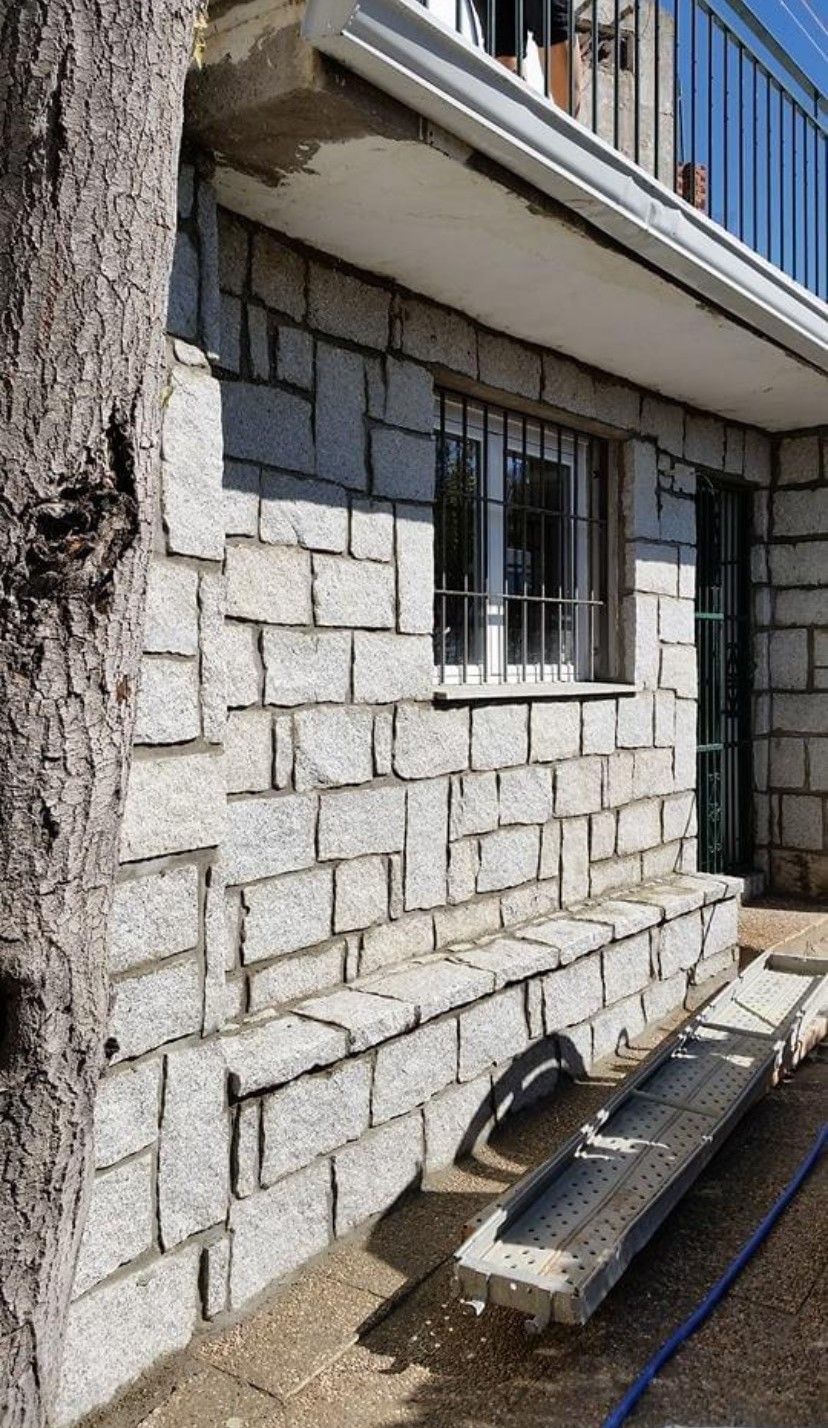Muro exterior de piedra con una pequeña ventana enrejada y un banco de madera debajo, junto al tronco de un árbol.