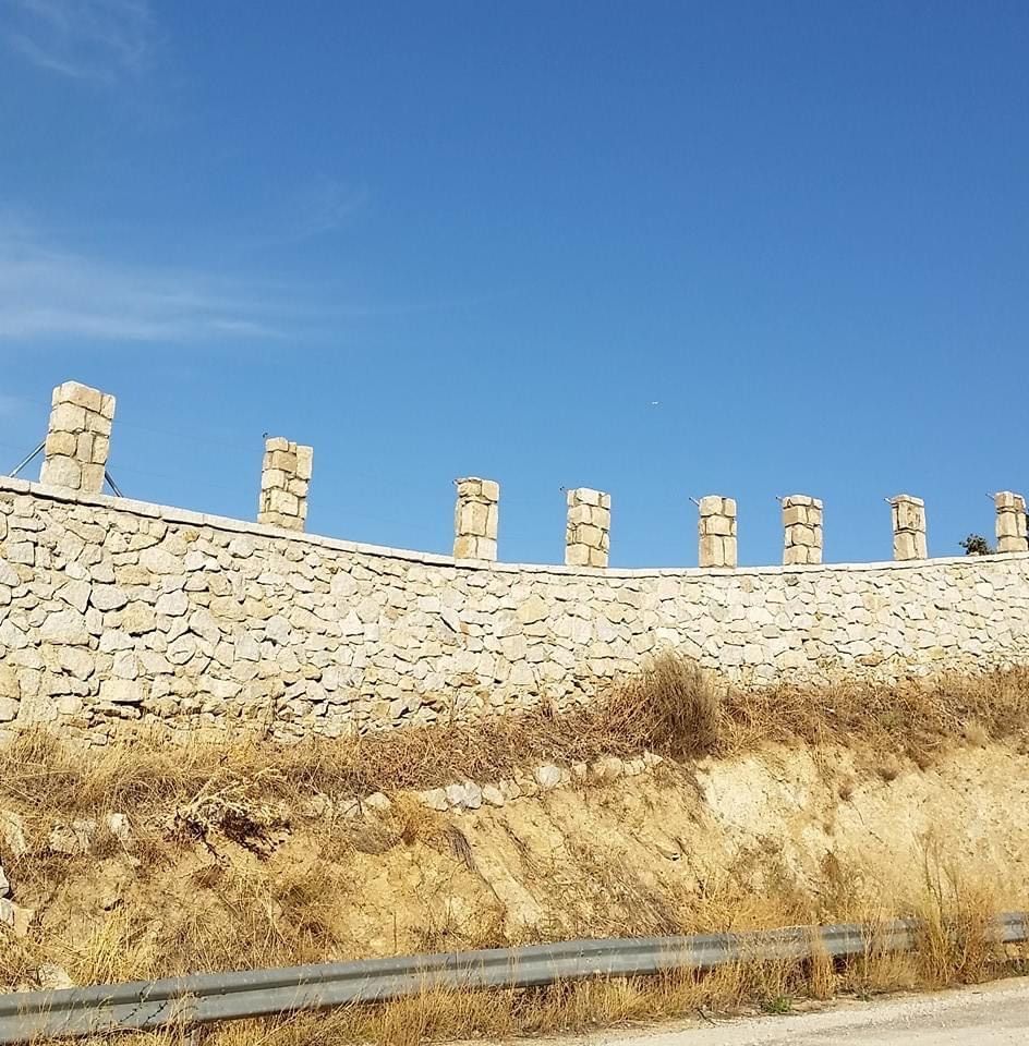 Muro de piedra con torres almenadas a lo largo de una carretera bajo un cielo azul despejado.