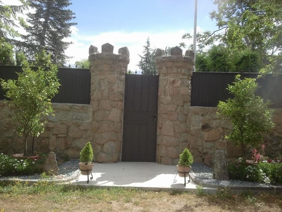 Muro de piedra en el jardín con una puerta central de madera, flanqueado por pilares y pequeños arbustos en la parte delantera.