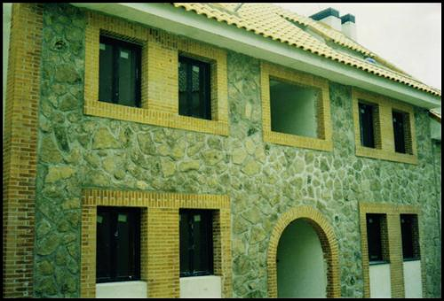 Fachada de casa de piedra con detalles amarillos, ventanas oscuras y una entrada arqueada.