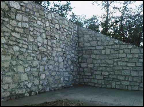 Esquina de muro de contención de piedra con suelo de hormigón y árboles visibles por encima.