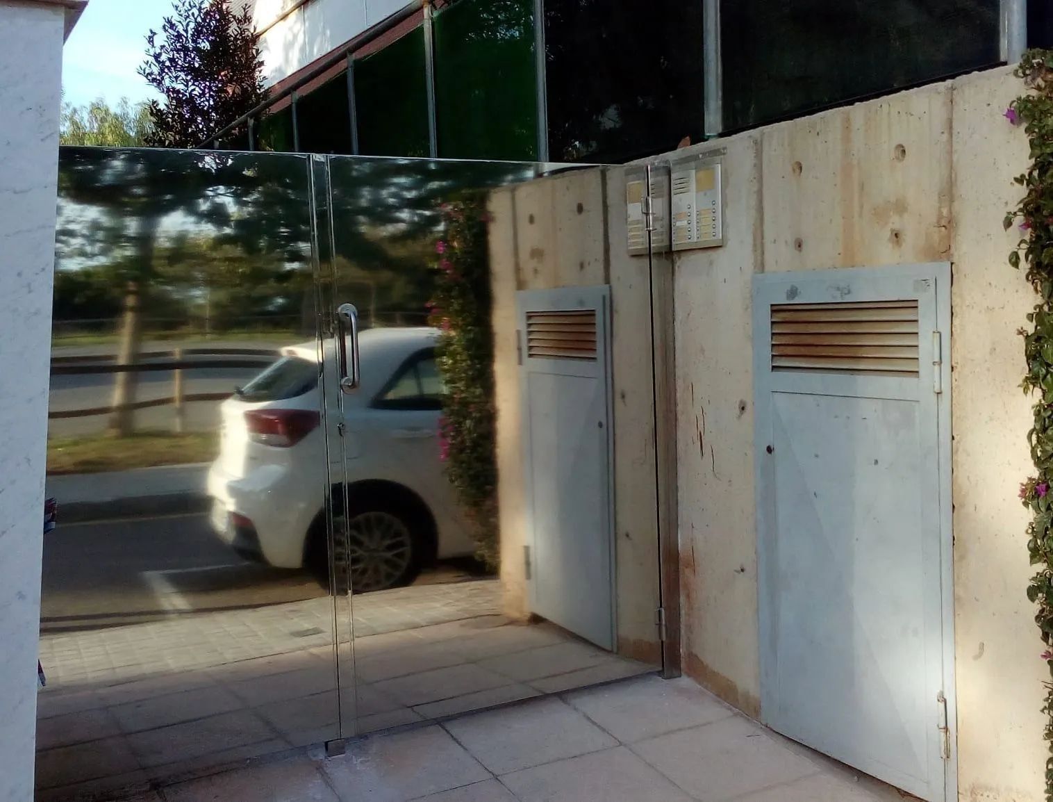Coche blanco aparcado cerca de una valla reflectante y dos puertas metálicas. Foto exterior, día soleado.
