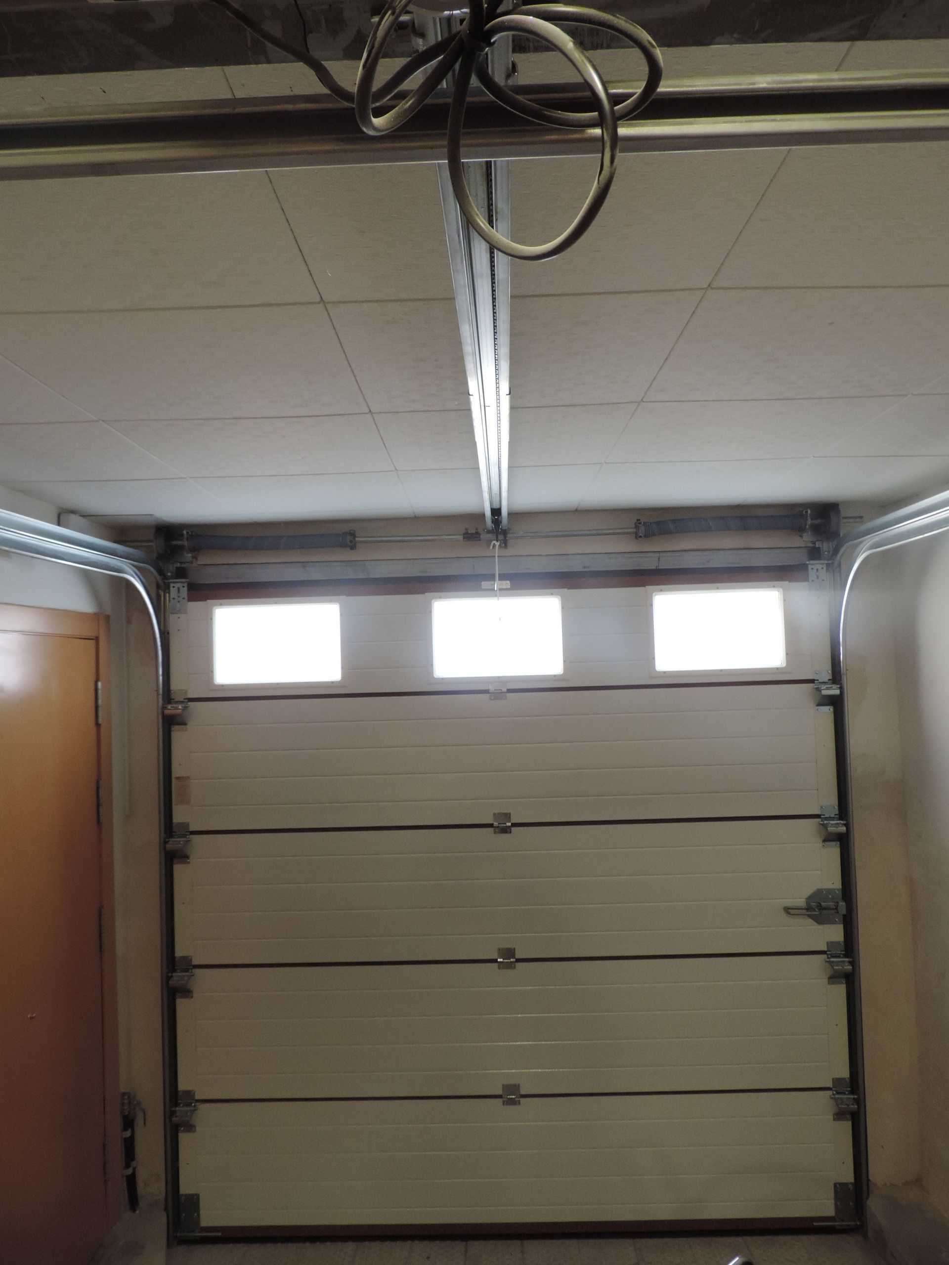 Puerta de garaje cerrada, con paneles horizontales. Riel de techo y mecanismo de apertura visibles.