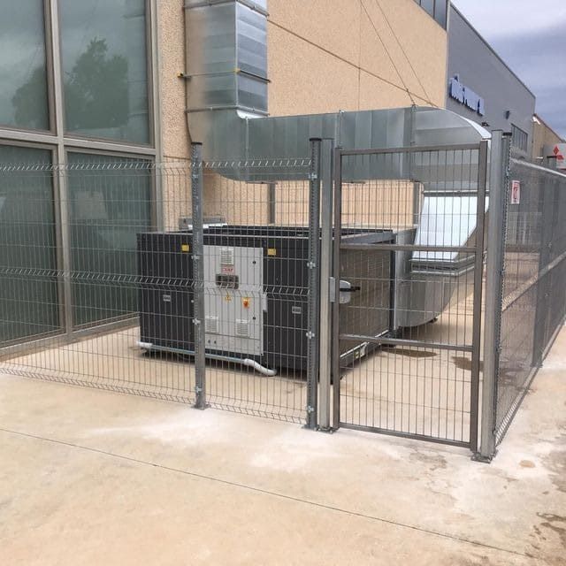 Una unidad HVAC industrial detrás de una valla de metal, al lado de un edificio con ventanas de vidrio.