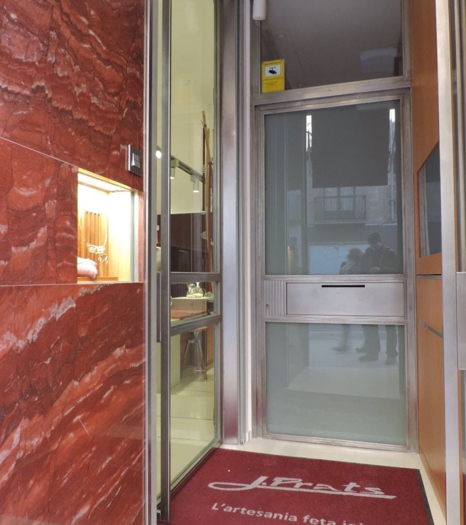 Entrada a una tienda con paredes de mármol rojo y puertas de cristal; se ve un felpudo rojo de bienvenida.