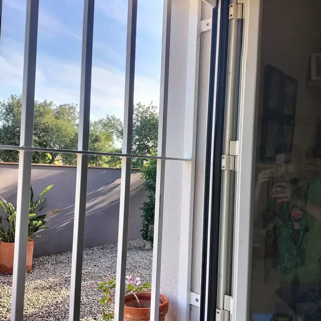 Vista a través de rejas de seguridad y una puerta de vidrio; patio exterior soleado con árboles y plantas.