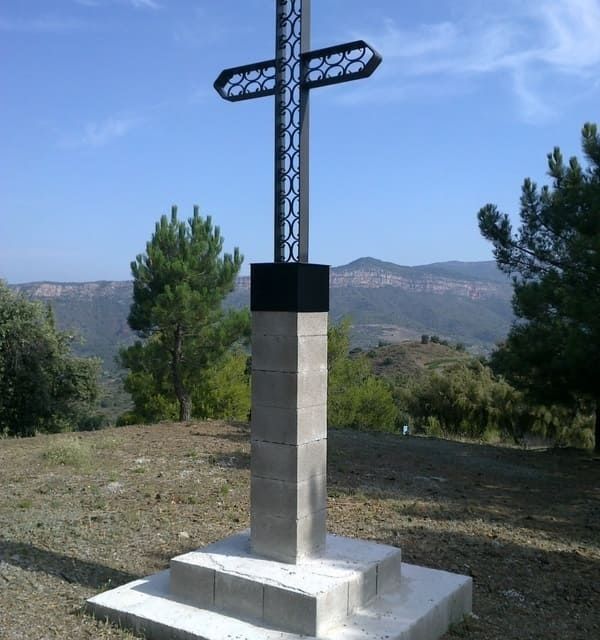 Cruz de metal sobre base de piedra, con un fondo de árboles y montañas bajo un cielo azul.