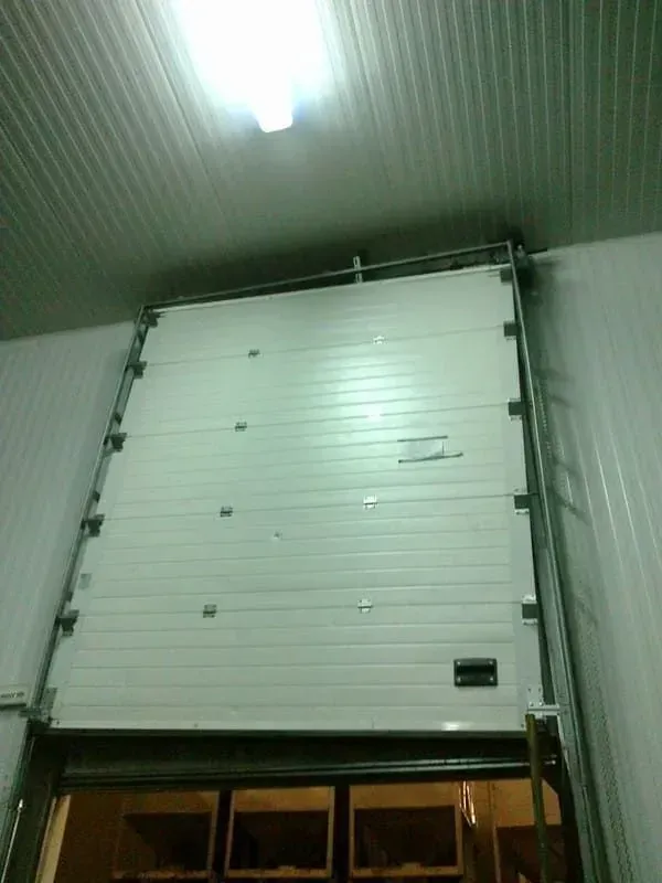 Puerta seccional blanca en un edificio, parcialmente abierta. Marco de acero, paneles horizontales, vista interior.