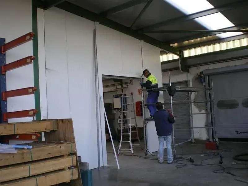 Trabajadores instalando paneles de pared en un almacén, uno de ellos utilizando una herramienta sobre una viga de metal.
