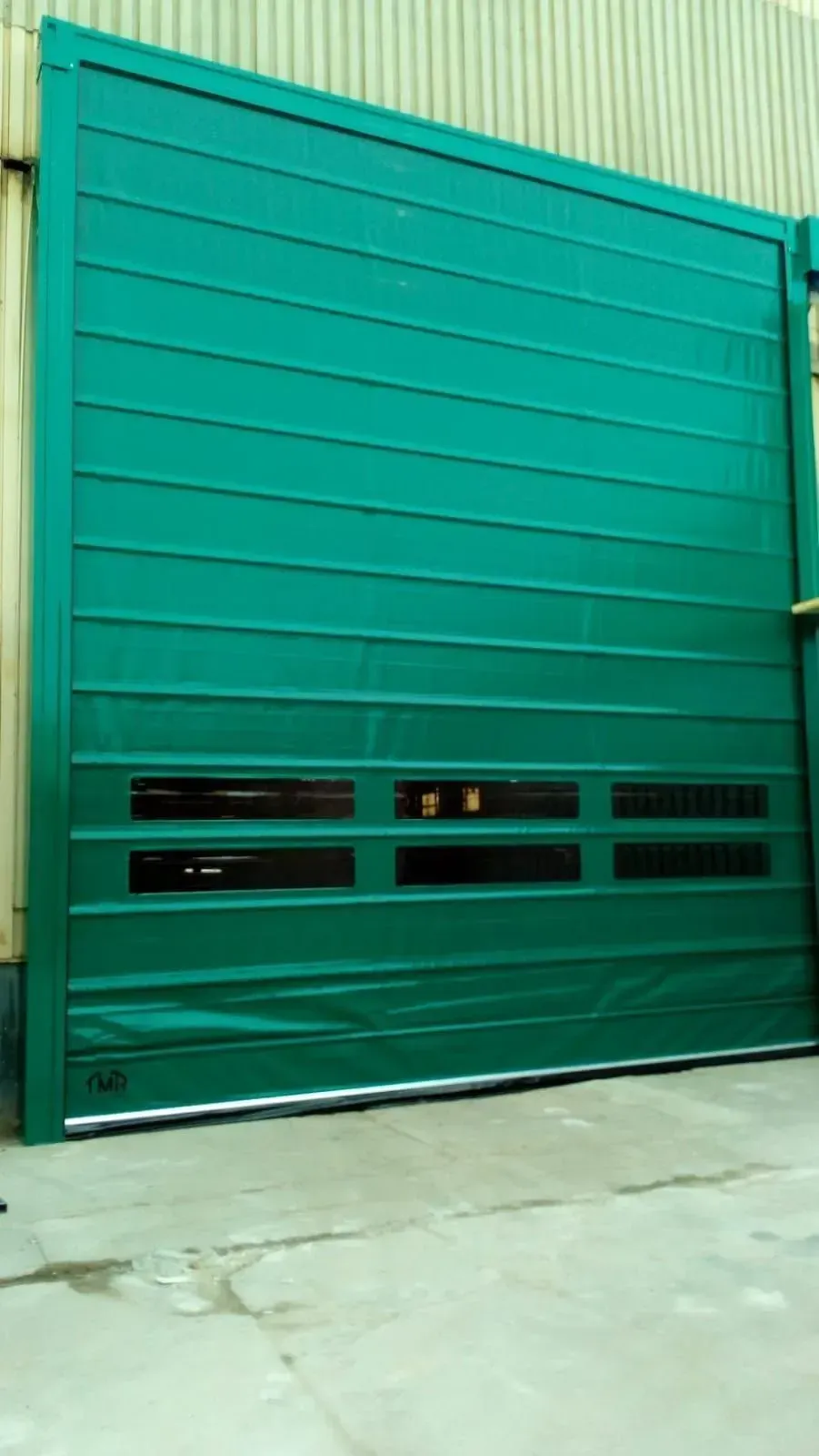 Puerta enrollable industrial de color verde con crestas horizontales, ventanas rectangulares y base metálica.