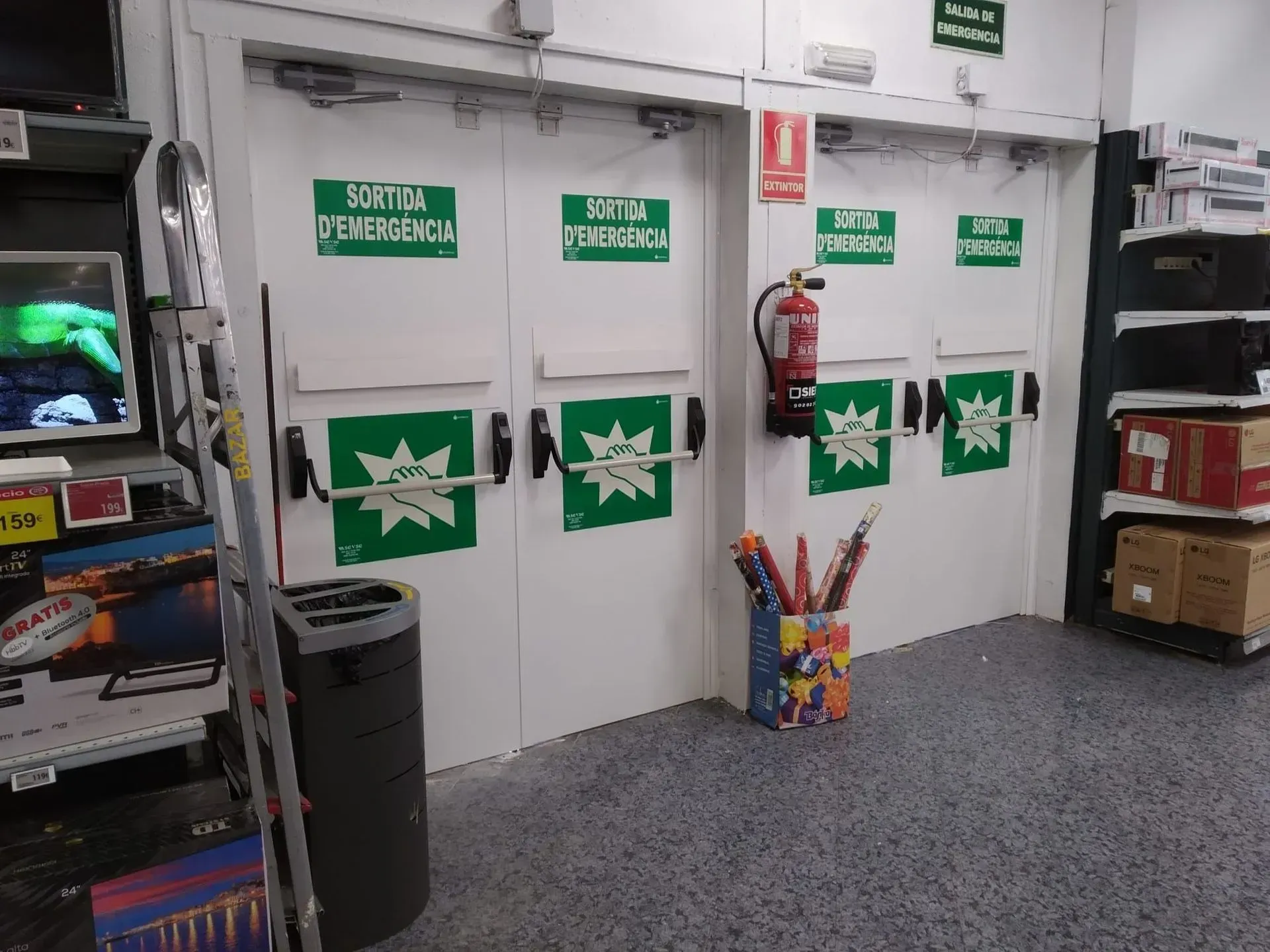 Tres puertas de salida de emergencia blancas con carteles verdes en una tienda, con extintor.