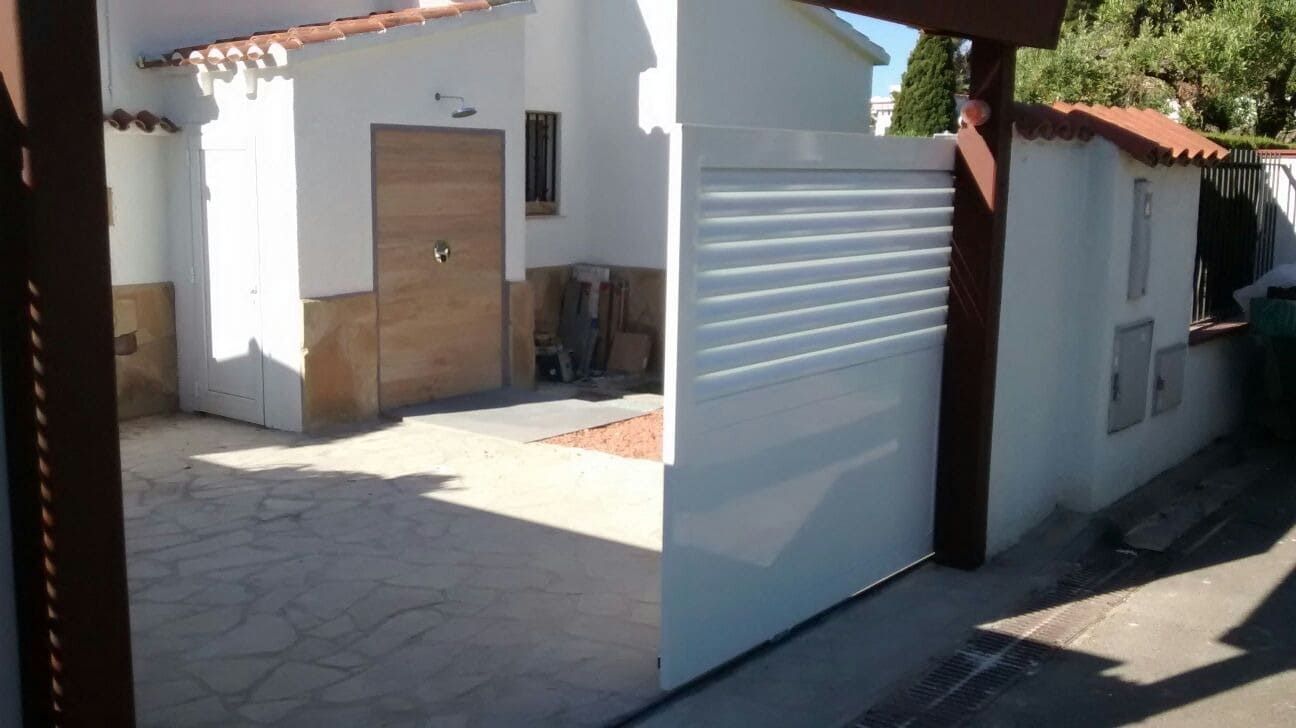 Entrada con portón blanco, puerta de madera y pequeño techo de tejas rojas.