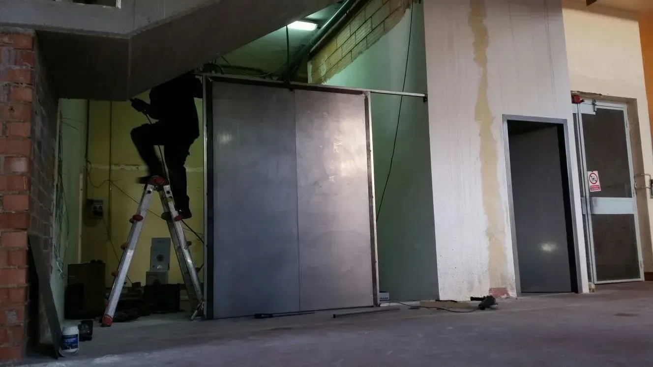 Persona en una escalera trabajando en grandes puertas metálicas dentro de un edificio.