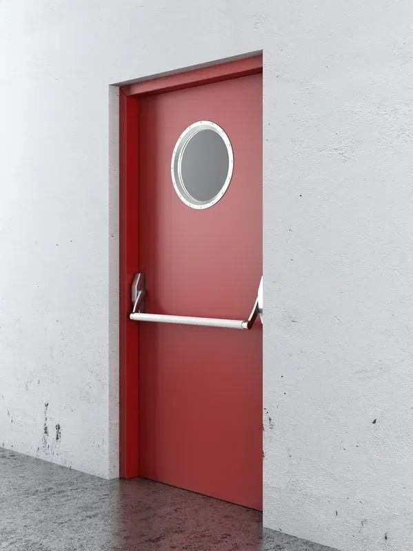 Puerta roja con ventana de ojo de buey y tirador plateado. Empotrada en pared blanca y suelo oscuro.
