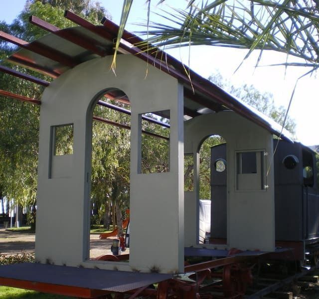 Vagón de tren parcialmente construido con aberturas arqueadas, ubicado en un parque. Construcción metálica gris y marrón.