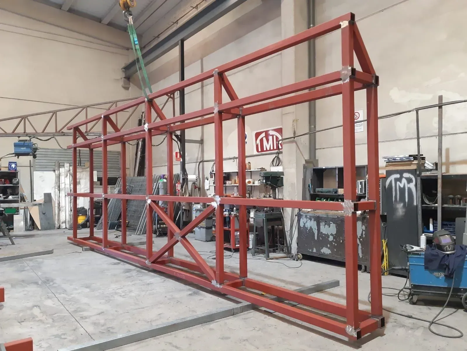 Construcción de estructura de acero en taller, pintada de rojo, con arriostramiento transversal y grúa.