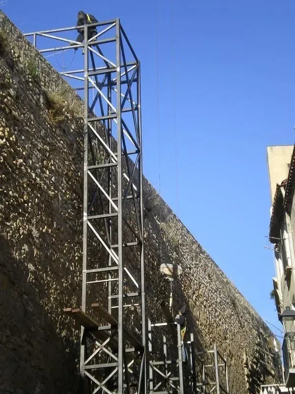 Andamio metálico junto a un alto muro de piedra, con un trabajador cerca de la cima, contra un cielo azul.