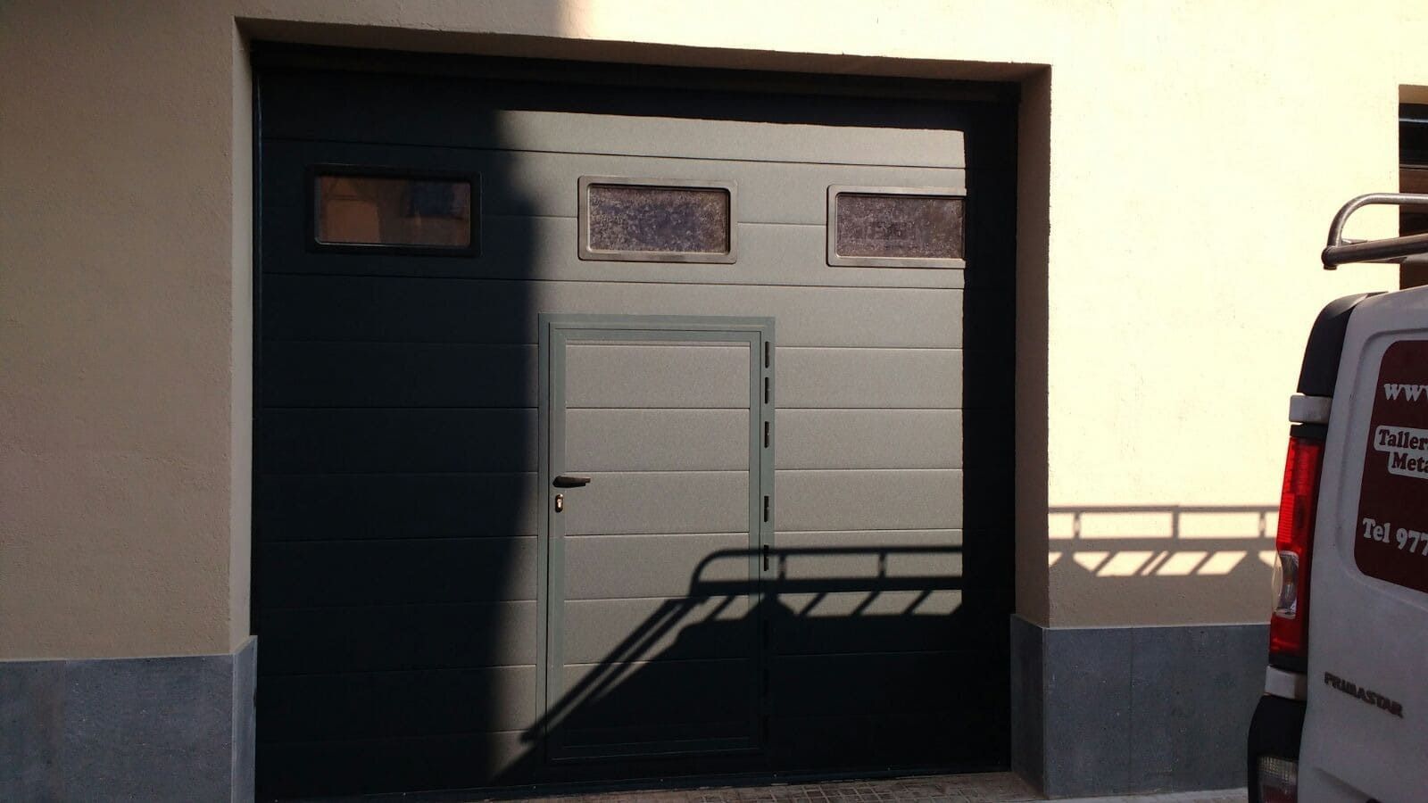 Puerta de garaje gris oscuro con una puerta pequeña, tres ventanas y una camioneta a la derecha.
