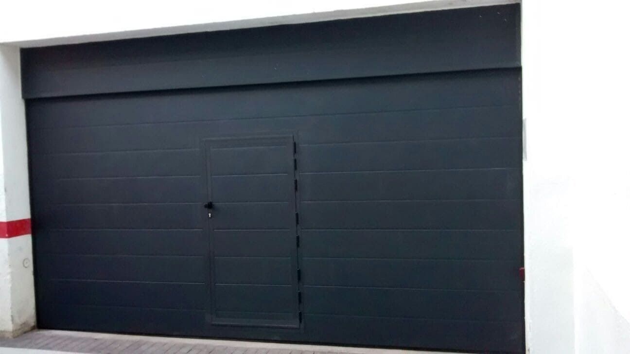 Puerta de garaje negra con un inserto de puerta más pequeño, colocada en una pared blanca.