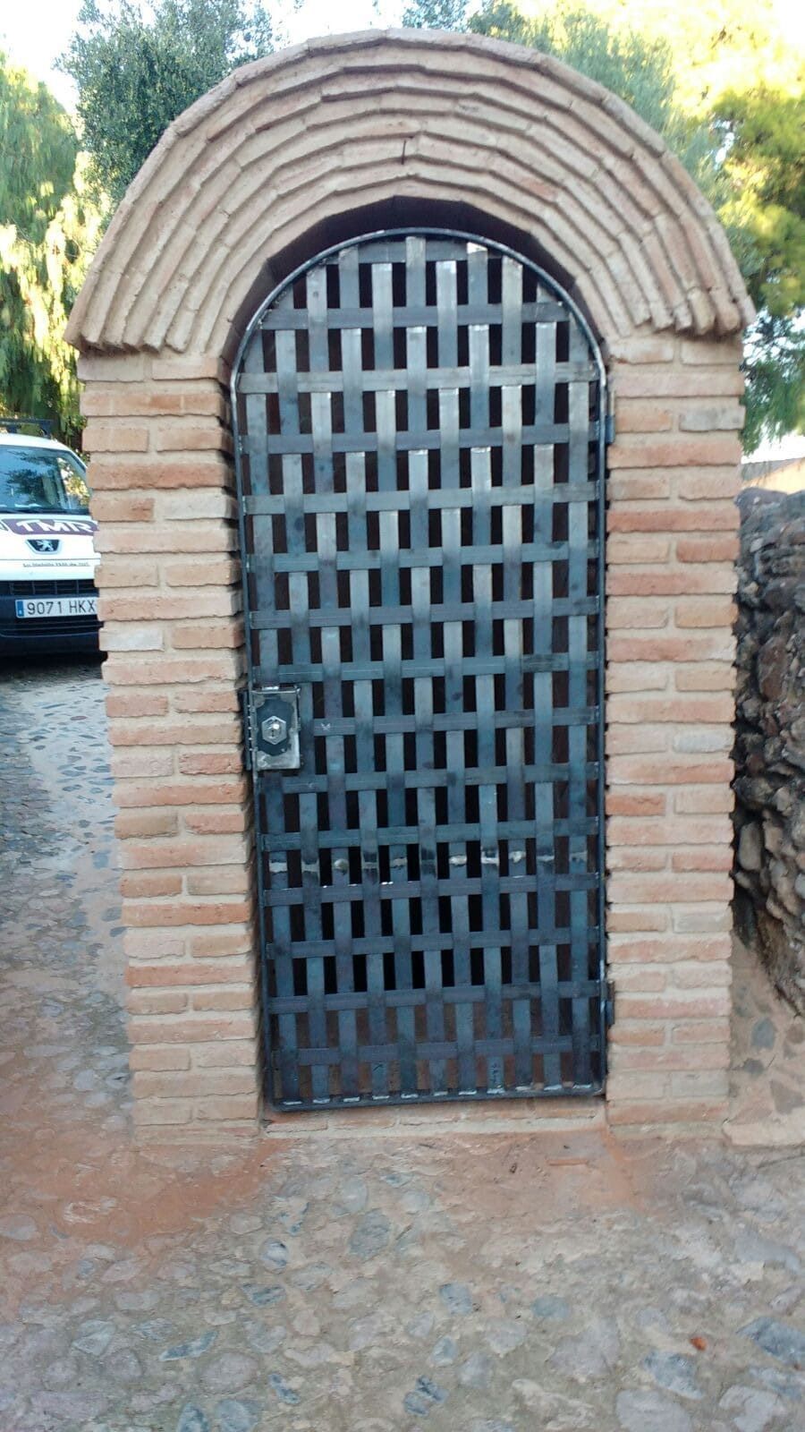 Arco de ladrillo con puerta metálica. Se ve parcialmente una furgoneta blanca a la izquierda.