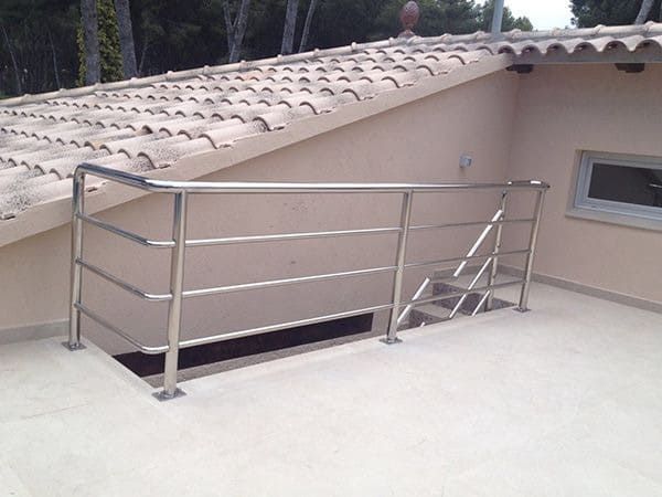 Barandilla de acero inoxidable sobre un tejado plano con una pequeña escalera de acceso. Paredes y tejas beige claro.
