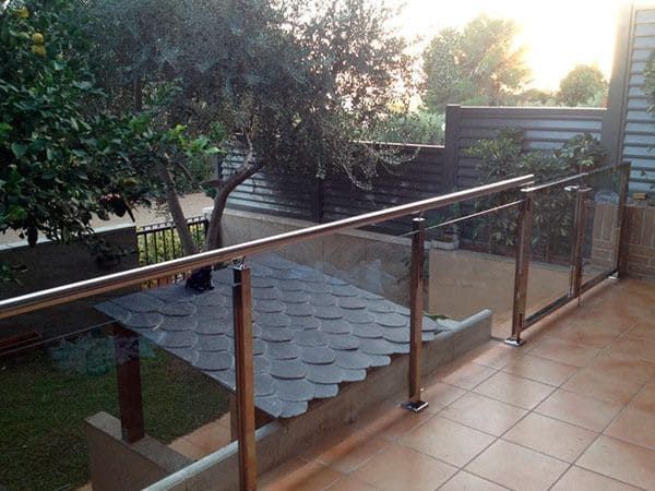 Un patio con barandilla de vidrio y metal, con vista a un patio con un árbol, escalones y un pequeño refugio.