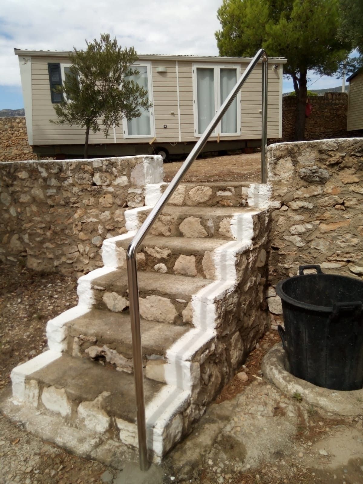Escalera de piedra con molduras blancas y pasamanos metálicos que conduce a una casa móvil. Cerca hay un cubo negro.