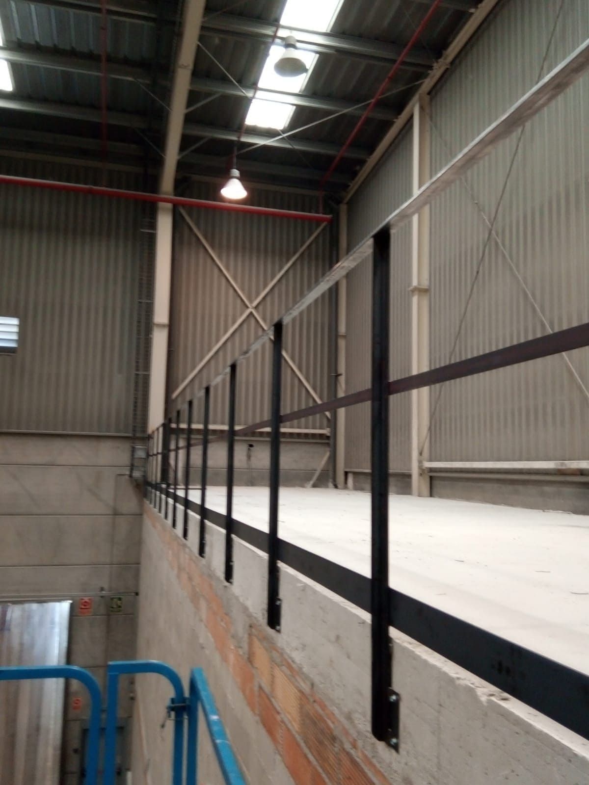 Pasamanos negro a lo largo de una pasarela de hormigón en el interior de un almacén con paredes de metal corrugado.