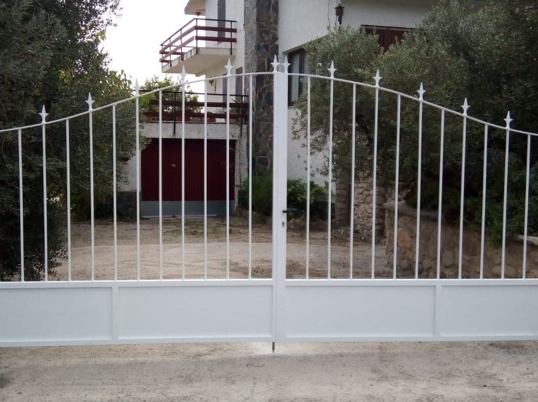 Portón metálico de color blanco frente a una casa, con rejas verticales y parte superior arqueada.