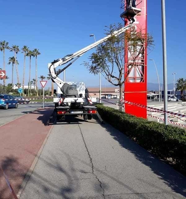 Un elevador montado en un camión recorta la vegetación alrededor de un cartel rojo alto en un día soleado.