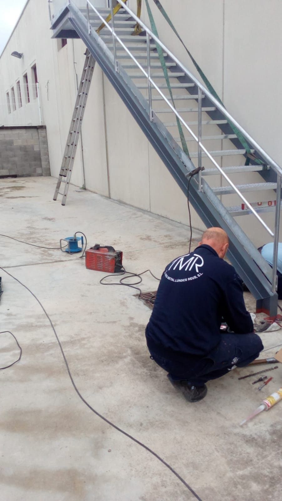 Un hombre trabaja en una escalera metálica de un edificio. Hay herramientas y una escalera cerca.