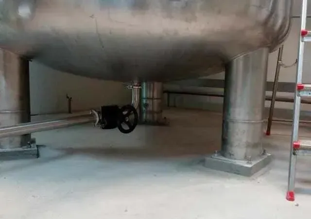 Gran tanque de acero inoxidable con tuberías y válvula en una habitación, sostenido por pilares, al lado de una escalera.