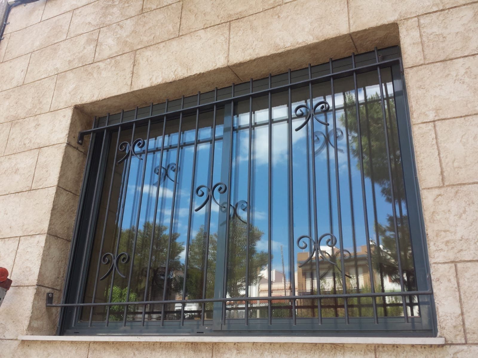 Ventana con rejas de seguridad negras que reflejan un cielo azul y árboles, situada en una pared de piedra clara.