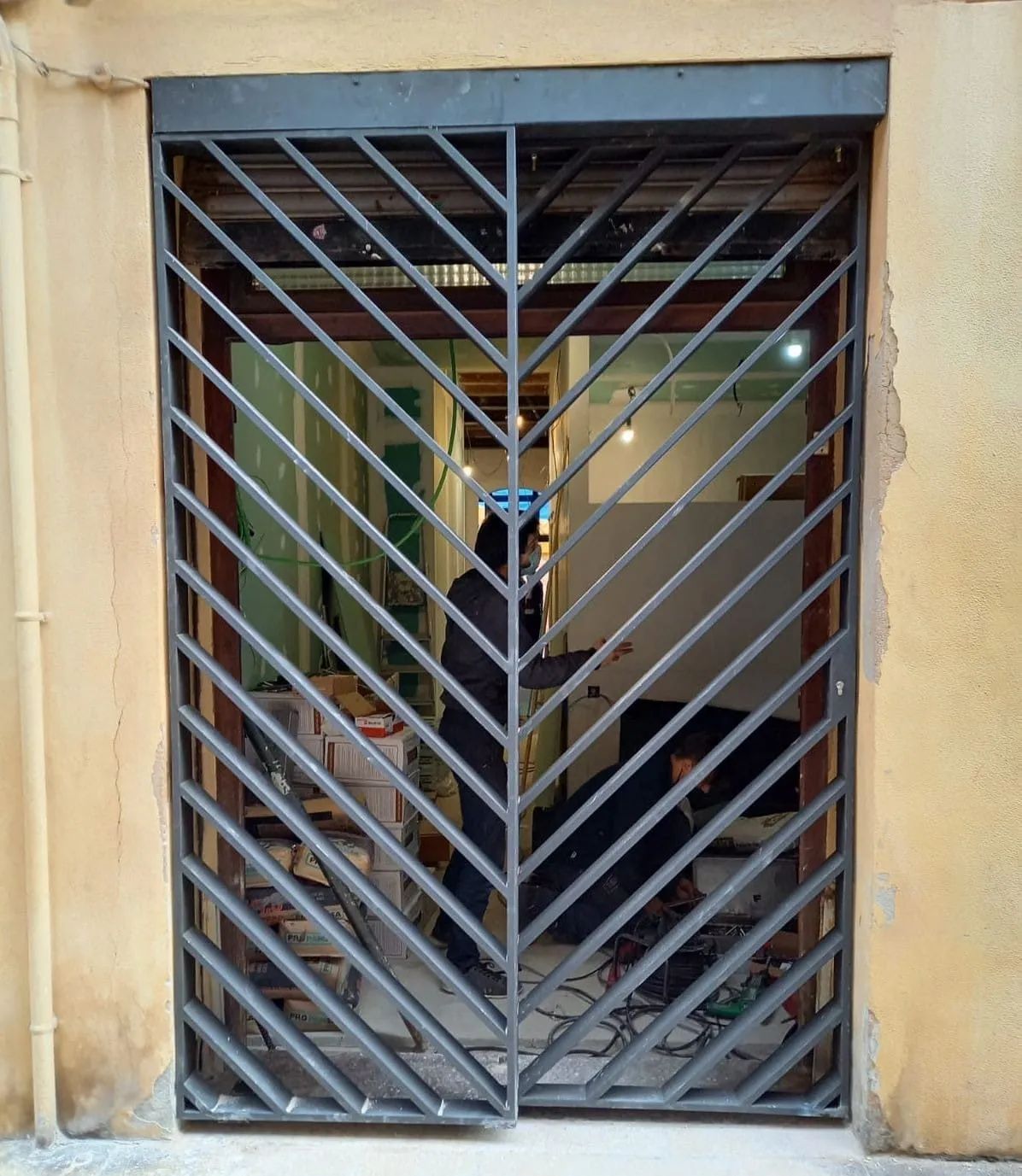 Puerta metálica con diseño de chevrones que cubre una entrada. Tras la puerta hay una sala con luces y algunos equipos.