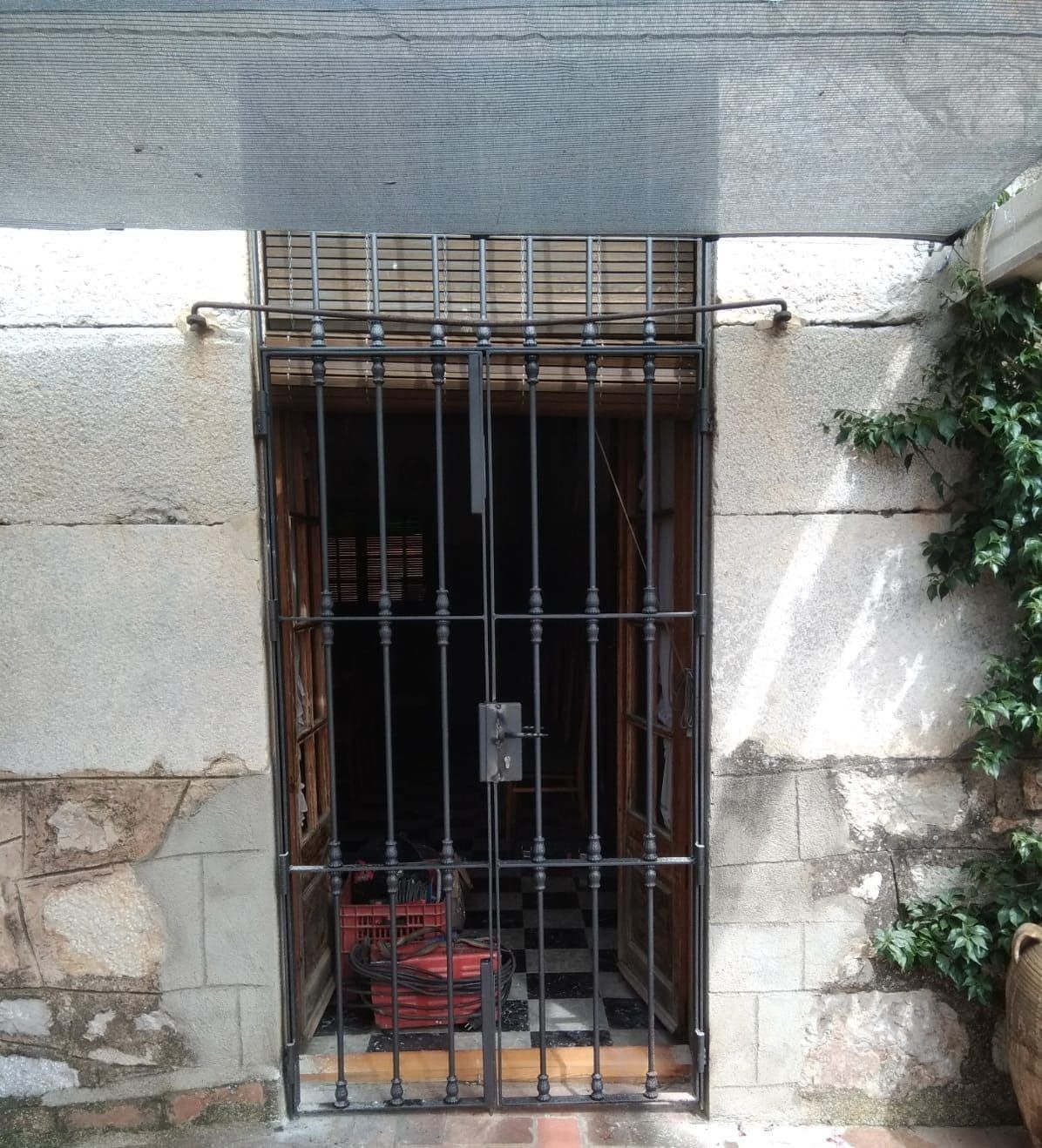 Puerta exterior con barrotes negros, carrito rojo en el interior. Muro de piedra beige y hiedra verde.