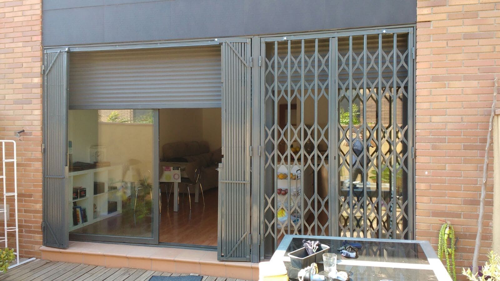 Puerta de seguridad de acordeón negra parcialmente abierta sobre puerta corrediza de vidrio, edificio de ladrillo.