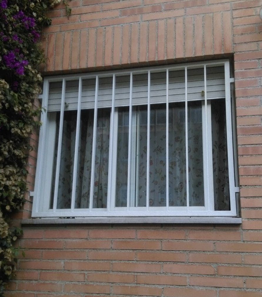 Ventana con barrotes blancos en un edificio de ladrillo con cortinas visibles en el interior.