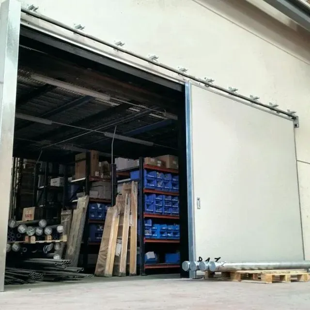Puerta industrial corrediza que se abre para revelar un área de almacenamiento con estantes, paletas y materiales metálicos.