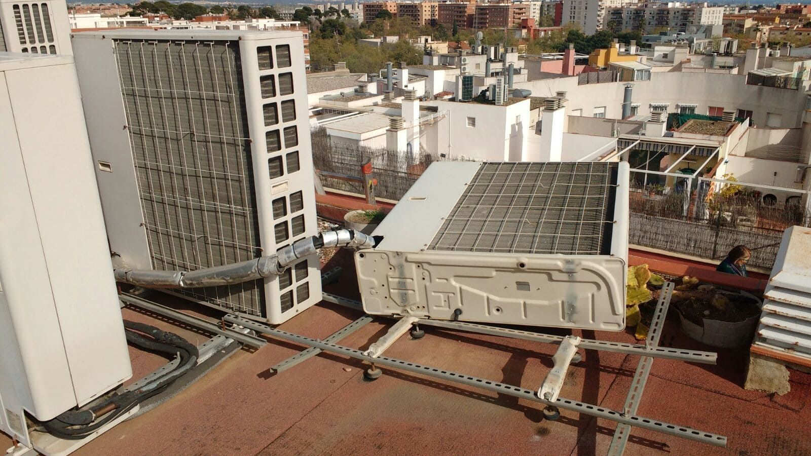 Azotea con dos grandes unidades de aire acondicionado, soportes metálicos y tuberías, sobre un fondo de paisaje urbano.